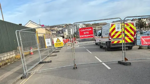 Vanessa Thomas A sign on steel gates reads Caution Site Entrance while a closed road in the background can be seen scattered with work site equipment and construction vehicles. 