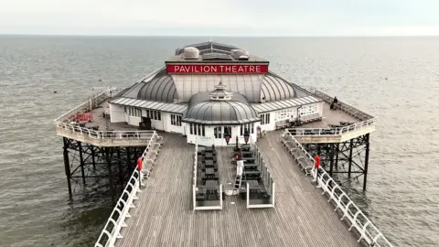 Shaun Whitmore/BBC A long wooden pier extends over calm sea water toward a building labelled “Pavilion Theatre.” White railings line both sides of the pier - with benches on the decking.