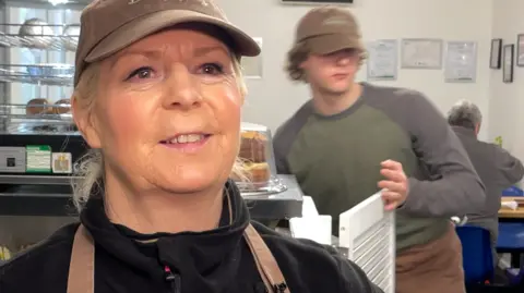 A blonde woman wearing a brown baseball cap and black top in a cafe and behind her a counter and a young man wearing a green jumper and brown trousers leaning on the counter