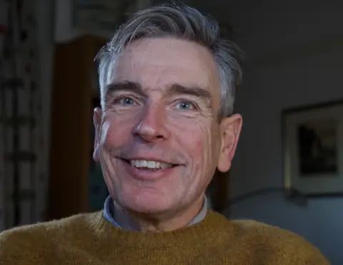 Dr Simon Rudland A man with grey hair smiles at the camera and is wearing a brown jumper