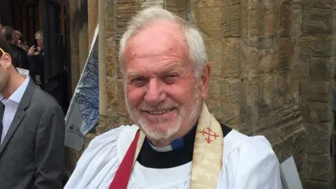 Family photo Reverend Gordon Driver in his robes after he was ordained in his sixties. He is smiling at the camera.