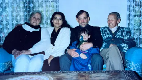 Serena Kern A family photo believed to have been taken around the year 2000 showing Carina Kern as a child with her parents and two grandparents. Four adults are sitting on a sofa (a man and woman in their 30s with dark hair in the centre, with an elderly parent on each side). On the left is Sydia Kern - she is around 70 years old, with grey hair tied back. She is smiling. Carina Kern is sitting on her father's knee. She is around six years old, wearing a blue tracksuit and holding a toy.