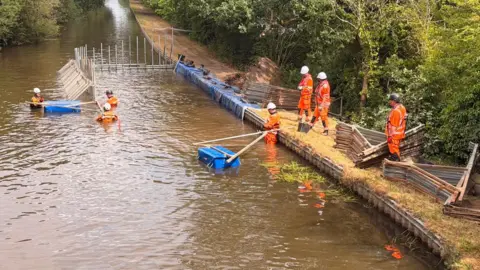 A section of canal, with workers standing on the towpath wearing orange high visibility clothing and other in the water. There is a metal structure in the water, which appears to be the framework of a temporary dam. There is blue tarpaulin on the side of the canal.