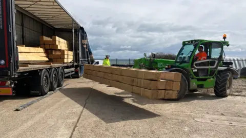 Environment Agency A green forklift truck lifts a number of large pieces of timber off the side of a lorry. 