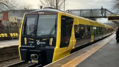 A yellow and black coloured Merseyrail train in a station. A person can be seen on the platform and another person is crossing the line on a bridge in the background