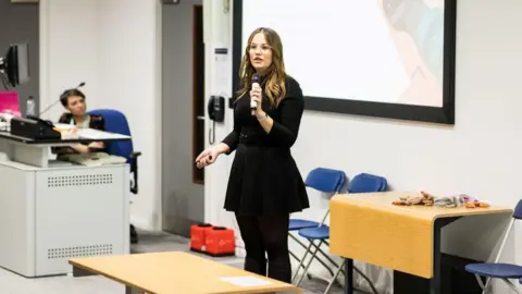 Reflect Digital A girl with long, brown hair wearing a black dress with black trousers. She is standing with a microphone at the front of a classroom with a screen behind her.