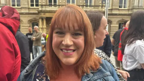 BBC/Crispin Rolfe Woman with shoulder length red hair, patterned top and denim jacket. She is stood in front of a crowd of people.