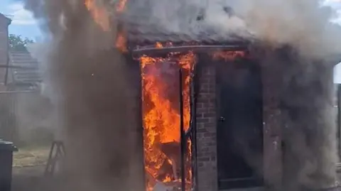 Robin Edmunds A red brick shed with two doorways with bright orange, fierce flames burning, one door destroyed so it can be seen. Thick black smoke surrounds it.