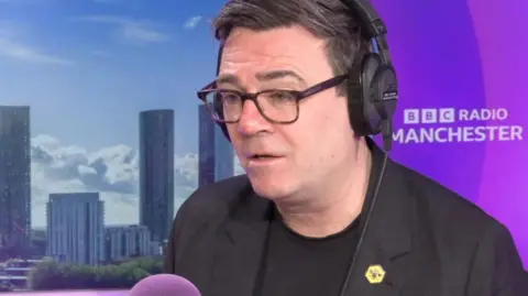 Andy Burnham is sitting in the BBC Radio Manchester studios. In the background are some skyscrapers from the city and the pink/purple local radio branding. Burnham has short dark hair and is wearing glasses, a dark jacket and dark top.