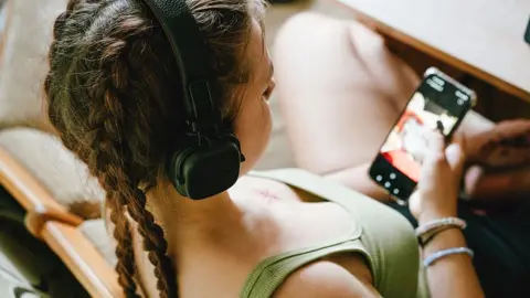 A close-up, over-the-shoulder stock photo shows a young person relaxing in a wooden chair while using a smartphone. The person wears large black over-ear headphones and has long hair styled in two braids. A light green sleeveless top is visible, along with bracelets on one wrist. The smartphone is held in one hand and displays a media or video-related screen, although the content is slightly out of focus. 