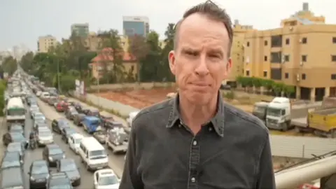 John Sudworth wearing a shirt with the top button undone. Behind him is a gridlocked road.