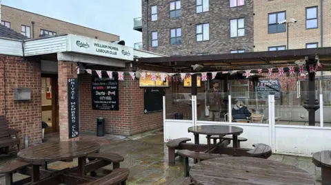 Martin Heath/BBC Entrance to brick-built club with William Morris Labour Club sign over the door (black writing on white background). The exterior includes a covered area containing tables, with a white wood and glass wall. There are brown tables with benches attached, resting on paving slabs.