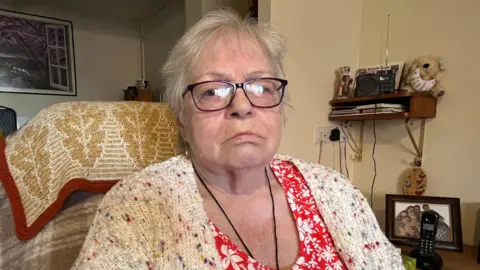 An elderly woman sits in an armchair and looks at the camera. She is not smiling. She wears a white cardigan with multi-colour spots knitted into it, a red blouse underneath with white flowers on it and glasses.
