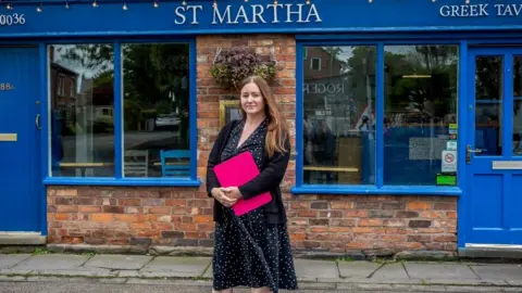 Kelly Woodnutt Kelly Woodnutt stood in front of her restaurant St Martha. She is wearing a dark polka dot dress and holding a pink clipboard. Her restaurant is blue.