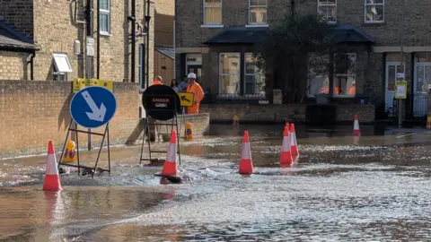 Jo Gee Water gushing out of an apparent hole on a road in Oxford. There are cones and signs around the hole.