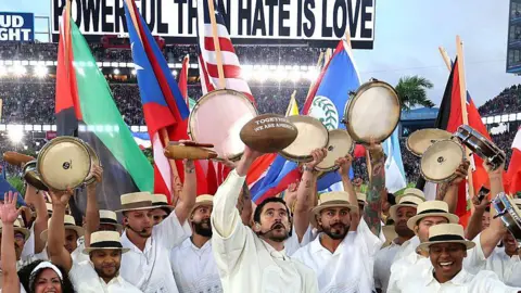 Getty Images Bad Bunny, surrounded by dancers in white shirts and Panama hats, performs at the Apple Music Super Bowl LX Halftime Show at Levi's Stadium. He holds an American football with "together we are America" printed across it above his head, as a sea of colourful flags rises behind him. 