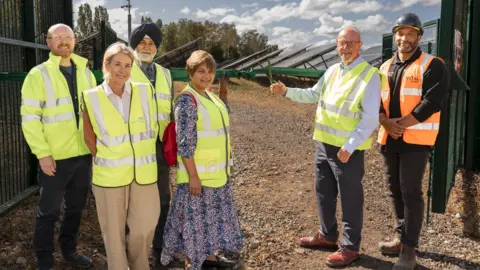 The Royal Wolverhampton NHS Trust Six people in hi-vis jackets are by a green ribbon and what appears to be an open green gate on the far right. Two men and two women are on the left and one of the two men on the right is holding scissors around the ribbon. 
