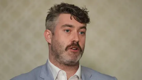 PA Media Eoin Ó Catháin is speaking to reporters in Washington. He has his mouth slightly open and is standing in front of cream, patterned wallpaper.