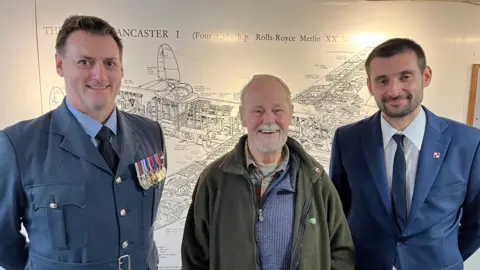 Three men face the camera, behind them is a picture of a Lancaster bomber, similar to the one which crashed 
