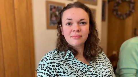 Amy smiles. She is sitting in a room with wooden furniture and blurred photos behind. She wears a polkadot blouse.