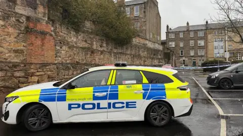 An Edinburgh street with parking spaces along the right. A police car is in the foreground with flats and police tape in the background. 