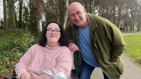 Iona Grant has dark hair down to her shoulders and is wearing a hair band and glasses. She is also wearing a pink woolly jumper. She is in a wheelchair in the outdoors. Next to her is her dad Stewart, who is wearing a green coat over a blue T-shirt. He is leaning on the back of her wheelchair There is grass and woodland behind them. They are both smiling at the camera.