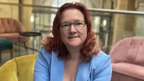 Kellie Coates. She is wearing a blue blazer, has shoulder-length red hair and is wearing glasses. She is sat on a yellow chair.