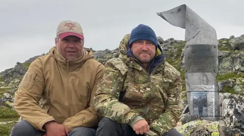 Espen Thronsen and Chris Bullock, sitting on rocky ground wearing outdoor weatherproof clothing and hats. Between them is a memorial made form World War Two aeroplane propellers. Attached to it in the middle is a plaque and beneath it is a photograph of Wilfred Surplice. 