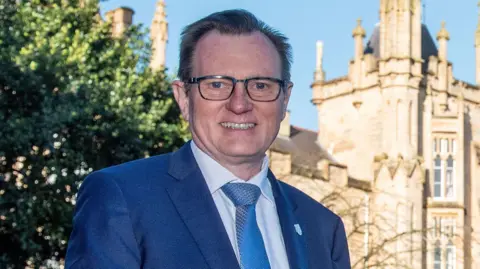 PA Media A man with dark hair, glasses and a blue suit with white shirt and blue tie smiling at the camera