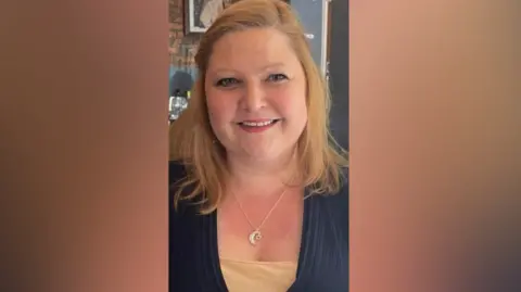 A woman smiles into the camera. She's in a restaurant. She's wearing a silver necklace with a small heart shaped dropping from a crescent moon and a black top over a beige vanity top. She has shoulder-length blond hair.