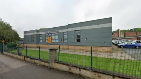 Google A grey single-storey school building behind a security fence 