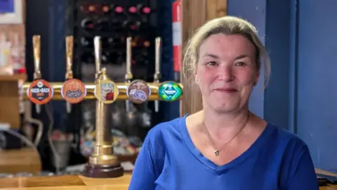 A wide shot of Fiona Hornsby standing in front of the bar at her pub. She has tied-back blonde hair and is wearing a blue top.