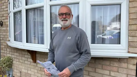 Tom Larsen-Wright/BBC A man in a grey sweatshirt and glasses stands in front of a bay window while holding some paperwork.