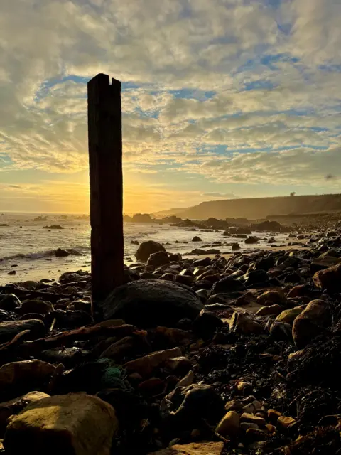 David Griffiths A golden sunset illuminates a rocky shoreline, with a single tall wooden post silhouetted against the sky.