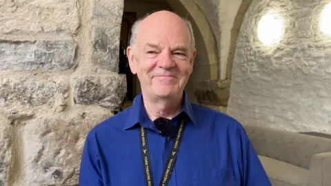 Kit Taylor/BBC Man with short grey hair, smiling and wearing a dark blue shirt and lanyard.