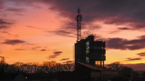The TT grandstand at sunset. The silhouette of the tower is set against an orange and pink sky. 