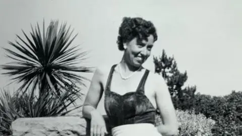 Black-and-white holiday photograph of Mrs Gay looking sideways and grinning while posing against a backdrop of palm trees and shrubbery.