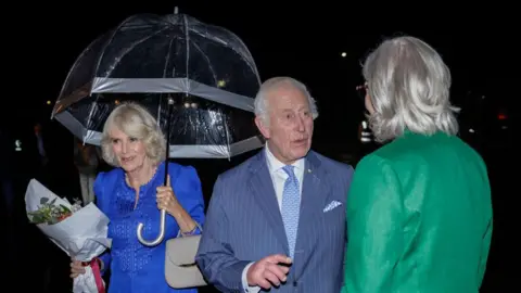 Brooke Mitchell / Getty Images Koning Charles III spreekt met de Australische gouverneur-generaal Sam Mostyn, terwijl koningin Camilla achter hem glimlachend een bos bloemen vasthoudt 