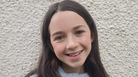 Rhea Flood standing against a white wall. She has long dark hair and brown eyes.