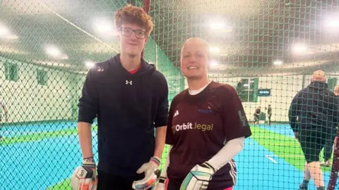 Jackie Storer A teenage boy and a woman are standing in an indoor cricket facility dressed in sports gear and cricket gloves. The boy has curly ginger hair and glasses, the woman has no hair.