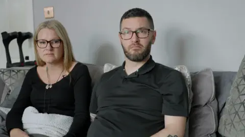 Redcar residents Clare and Paul, who were affected by the cyber-attack. They are sitting on a grey sofa covered in cushions. Both wear black tops and dark rimmed glasses. Clare has shoulder length blonde hair with a centre parting. Paul has short black hair and a groomed beard and moustache. 