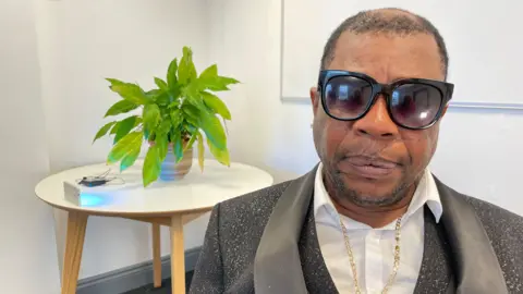 Annabel Amos/BBC Rohan Shand Snr wearing a grey suit, gold necklace and sunglasses. Behind him is a small round table with a green plant on top.