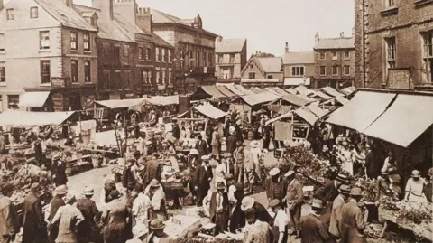 Isabel Garbutt Collection (donated to Knaresborough Museum Association) A black and white photo showing lots of people in the 1920s around many stalls in a market place