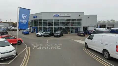 Google Bristol Street Motors showroom. A load of vehicles are parked beside a large grey building with large windows.