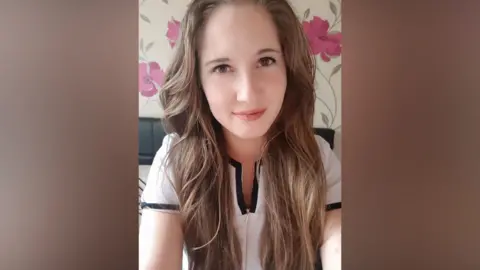 A young woman with long brown wavy hair is looking into the camera while sat in a bedroom. She's wearing a  short sleeved white blouse with a black lining. 