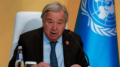 Reuters UN secretary general Antonio Guterres pictured during a speech. He is an elderly man with short, grey hair, combed to one side. He wears a grey suit jacket, a white shirt, and a patterned blue tie. He sits in front of a yellow wall with a bright blue UN banner beside him, speaking into a small black microphone.