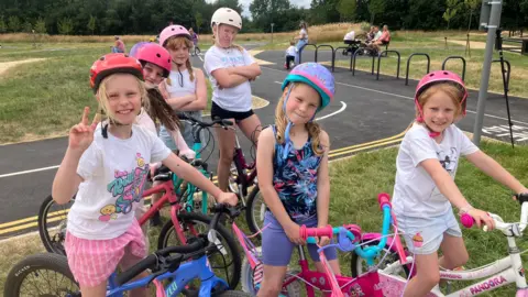 Six girls sit on bikes in a bike park. They have pink, white, purple and red helmets on. They are all seven years old.