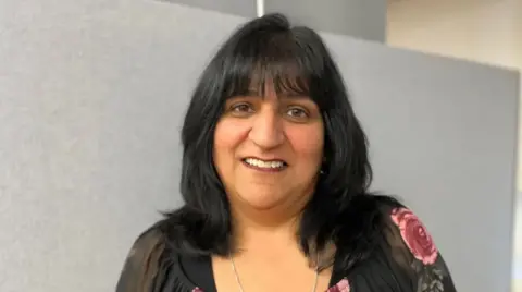 A woman with long black hair and a black top with a pink floral pattern on it in a room with a grey wall