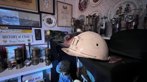 A white miner's helmet on a sideboard in a room, surrounded by lamps, pictures and signs from the coal mining industry.