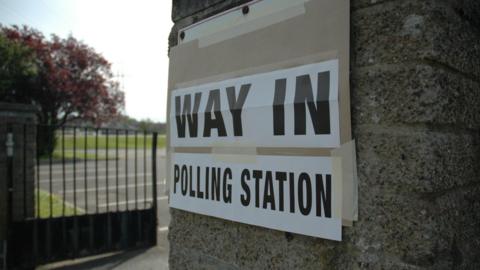 a sign saying Way in polling station. The sign is raped to a stone wall and a railing and some parkland can be seen in the background